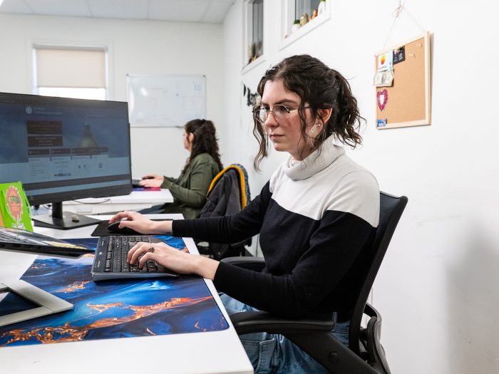 A woman with brown hair and glasses working at a computer in an office. There is another person working on a computer in the background.