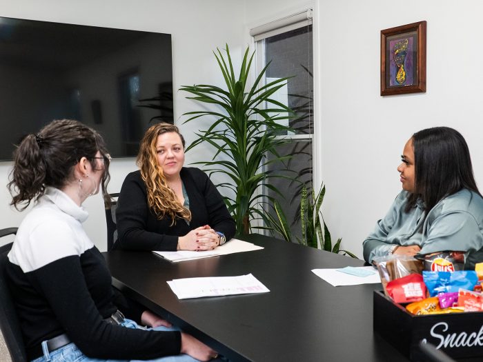Three women sitting around a conference table with papers and snacks in front of them.