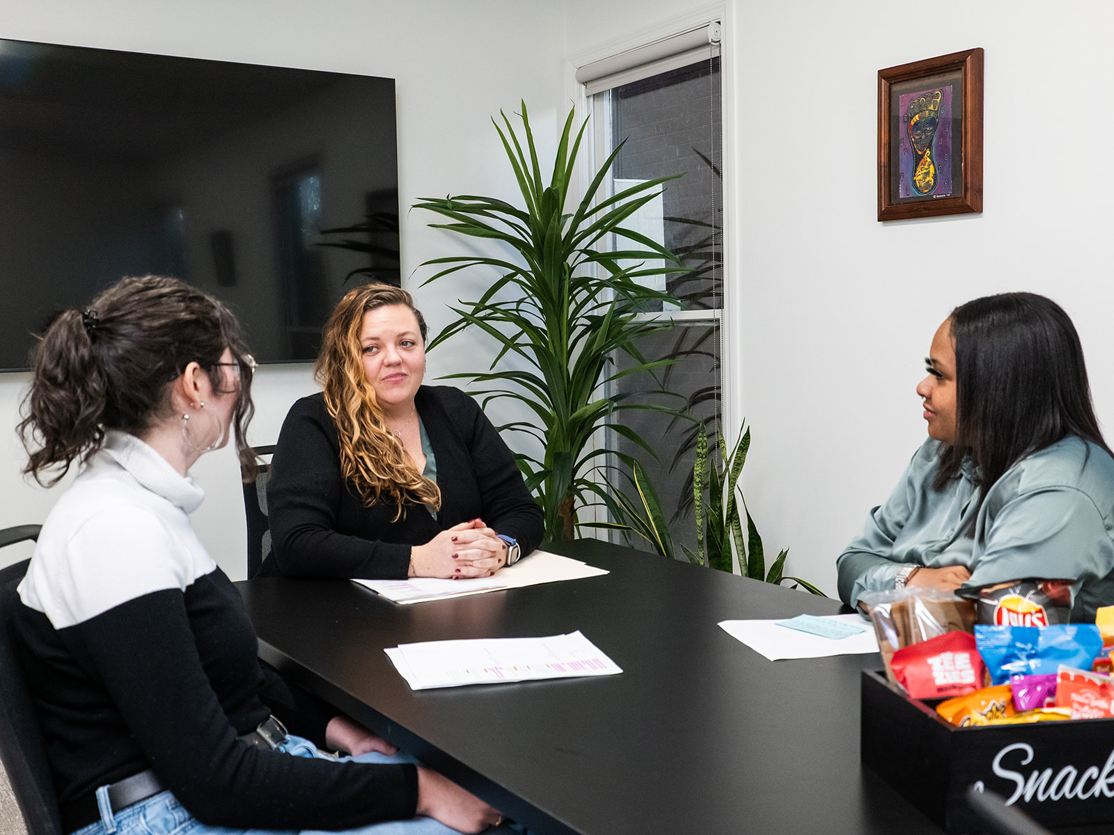 Three women sitting around a conference table with papers and snacks in front of them.