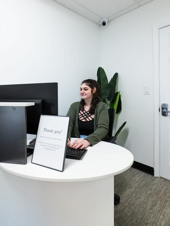 A dark-haired woman is typing at a desk behind a computer monitor and a sign that says "Thank you!"