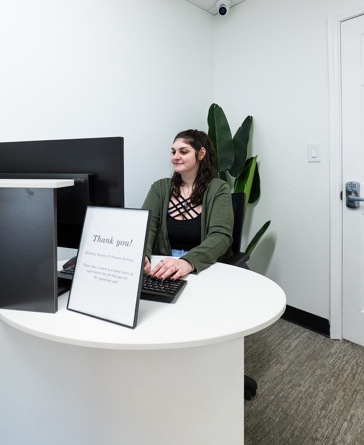A dark-haired woman is typing at a desk behind a computer monitor and a sign that says "Thank you!"